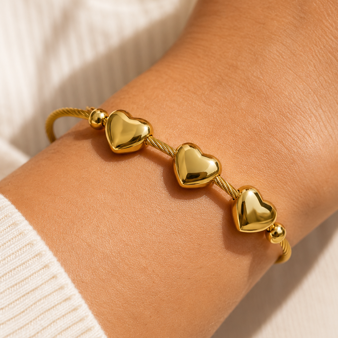 A close-up of a gold heart charm bracelet worn on a person's wrist, featuring three polished heart charms connected by twisted rope-style links on a white ribbed knit sleeve.