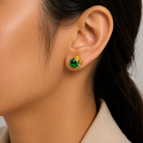 A close-up of a person's ear wearing a small gold stud earring with a round green faceted gemstone and tiny clear accent stones.