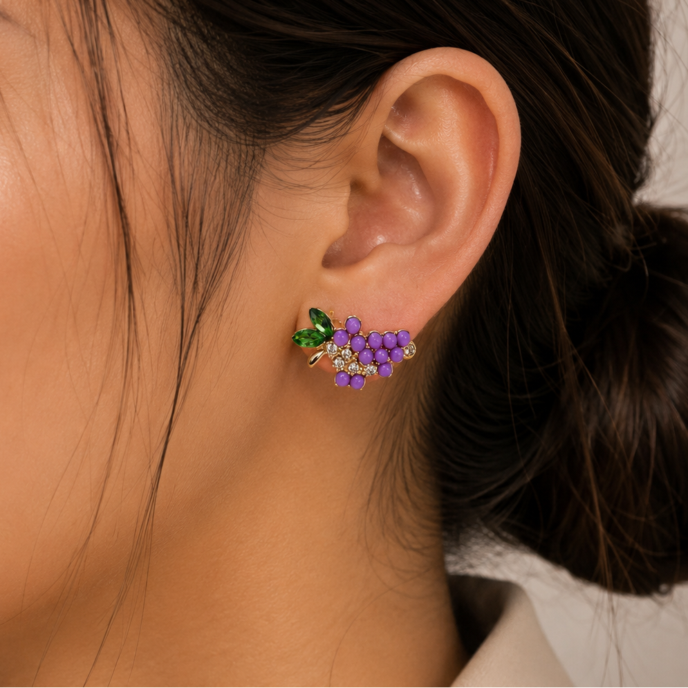 A close-up of a woman's ear wearing a small grape cluster stud earring with purple grapes, green leaves, and tiny clear stones.