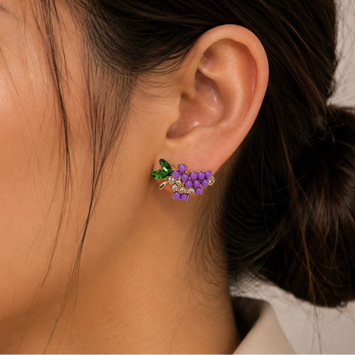 A close-up of a woman's ear wearing a small grape cluster stud earring with purple grapes, green leaves, and tiny clear stones.