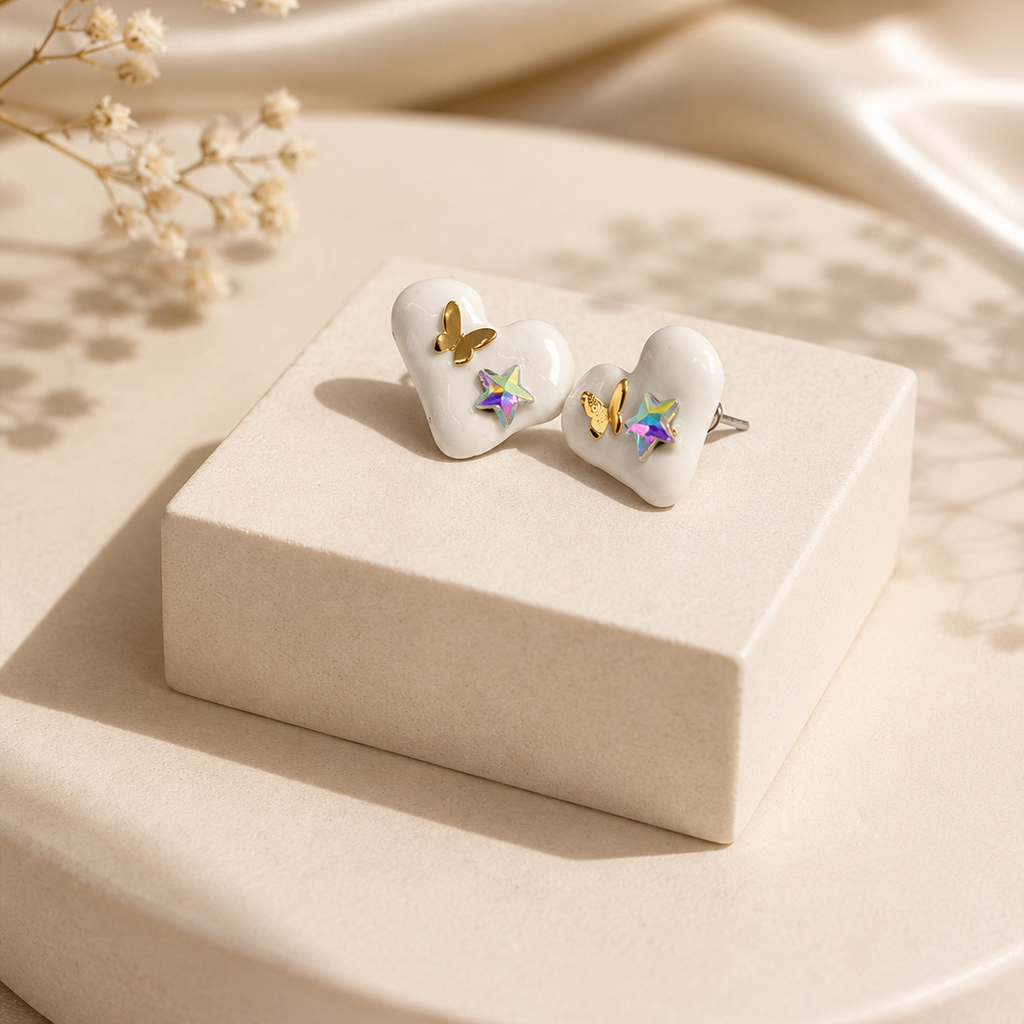 A pair of white heart-shaped stud earrings with small gold and iridescent butterfly details, displayed on a cream-colored rectangular box with dried baby's breath flowers and draped satin fabric in the background.