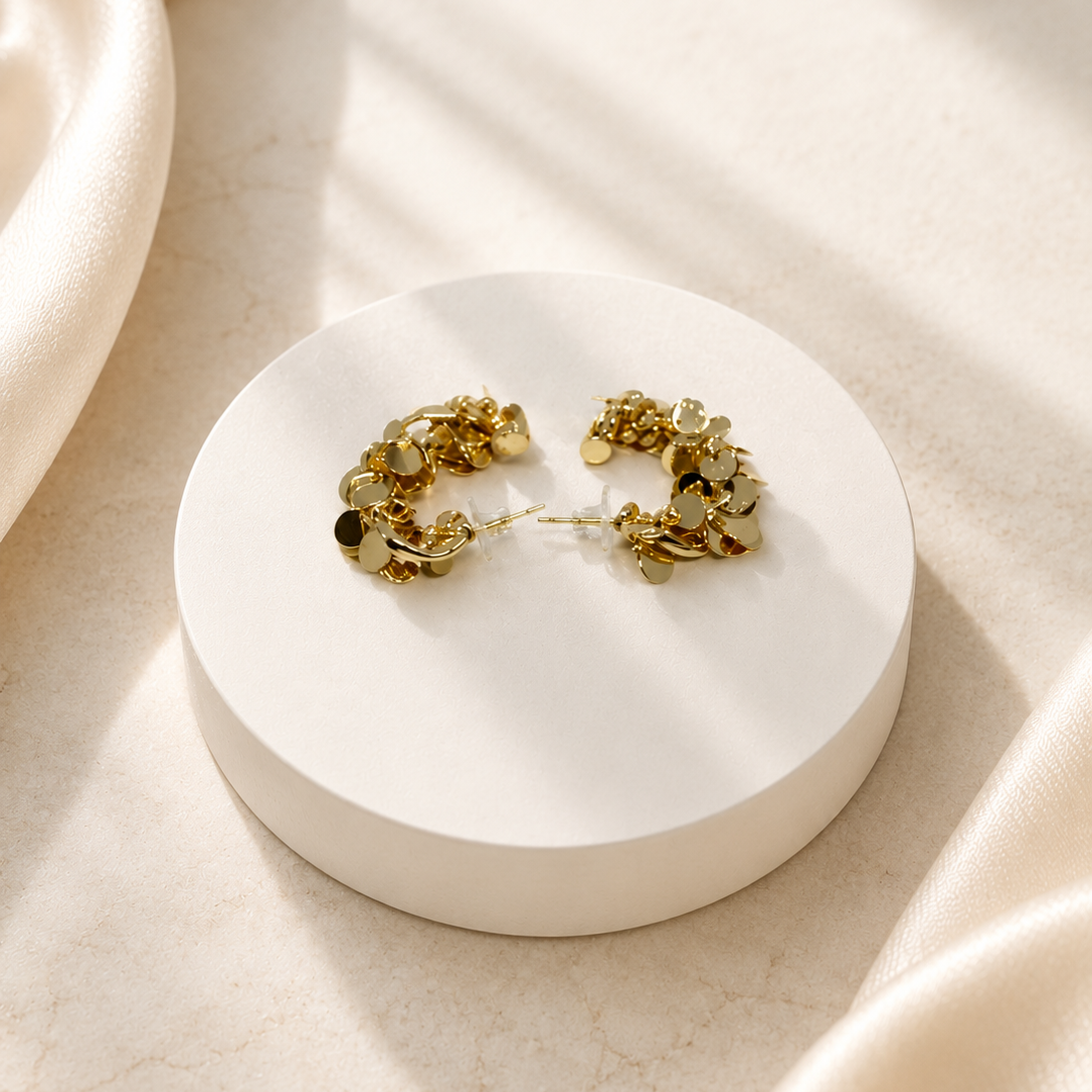 A pair of gold-toned floral hoop earrings displayed on a round white pedestal, set against a warm beige marble surface with draped cream fabric and soft window shadows.