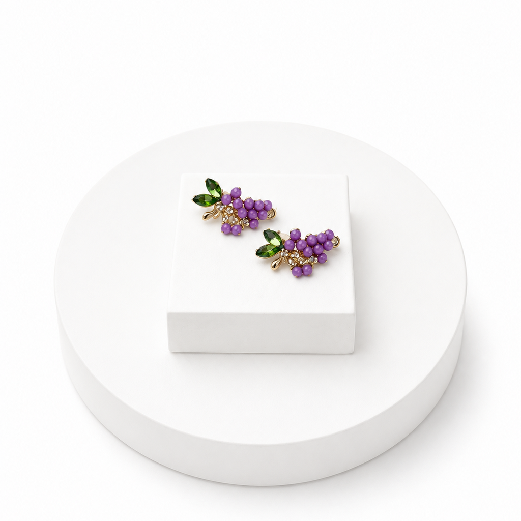A pair of grape cluster stud earrings with purple grapes, green leaves, and gold-tone metal accents, displayed on a small white block atop a round white pedestal against a white background.