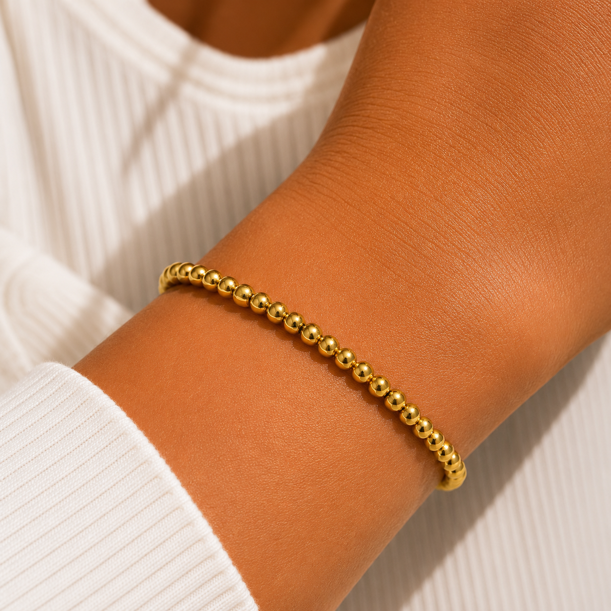 A delicate gold beaded bracelet worn on a person's wrist, shown against a white ribbed knit top.
