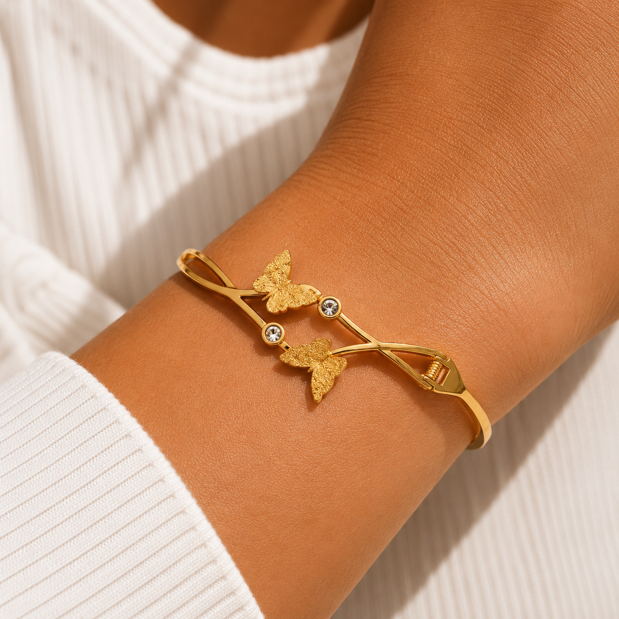 A gold-toned bangle bracelet with two butterfly charms and small clear stones, shown worn on a person's wrist over a white ribbed top.