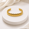 A shiny gold cuff bracelet with a row of teardrop-shaped links, displayed on a round white pedestal with soft white fabric in the background.