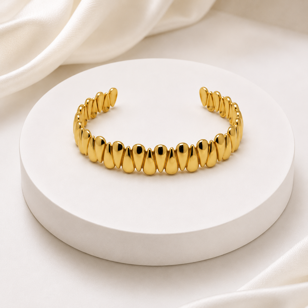 A shiny gold cuff bracelet with a row of teardrop-shaped links, displayed on a round white pedestal with soft white fabric in the background.