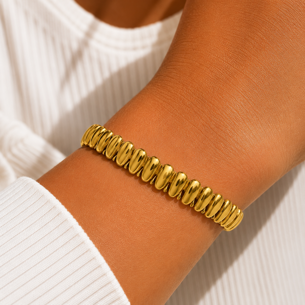 A close-up of a gold beaded stretch bracelet worn on a person's wrist, paired with a white ribbed long-sleeve top.