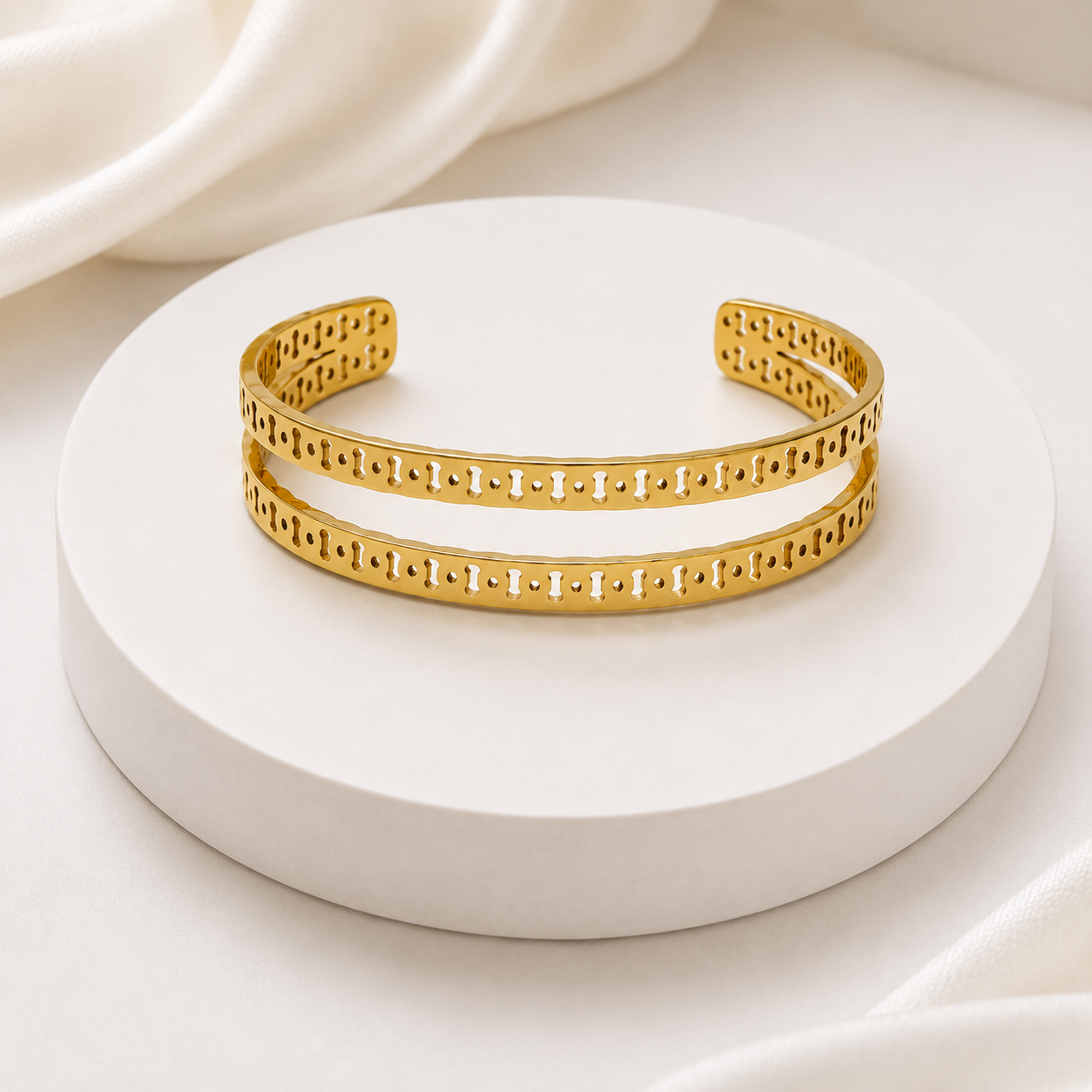 A pair of gold-plated open cuff bracelets with a detailed geometric cutout pattern, displayed on a round white pedestal.