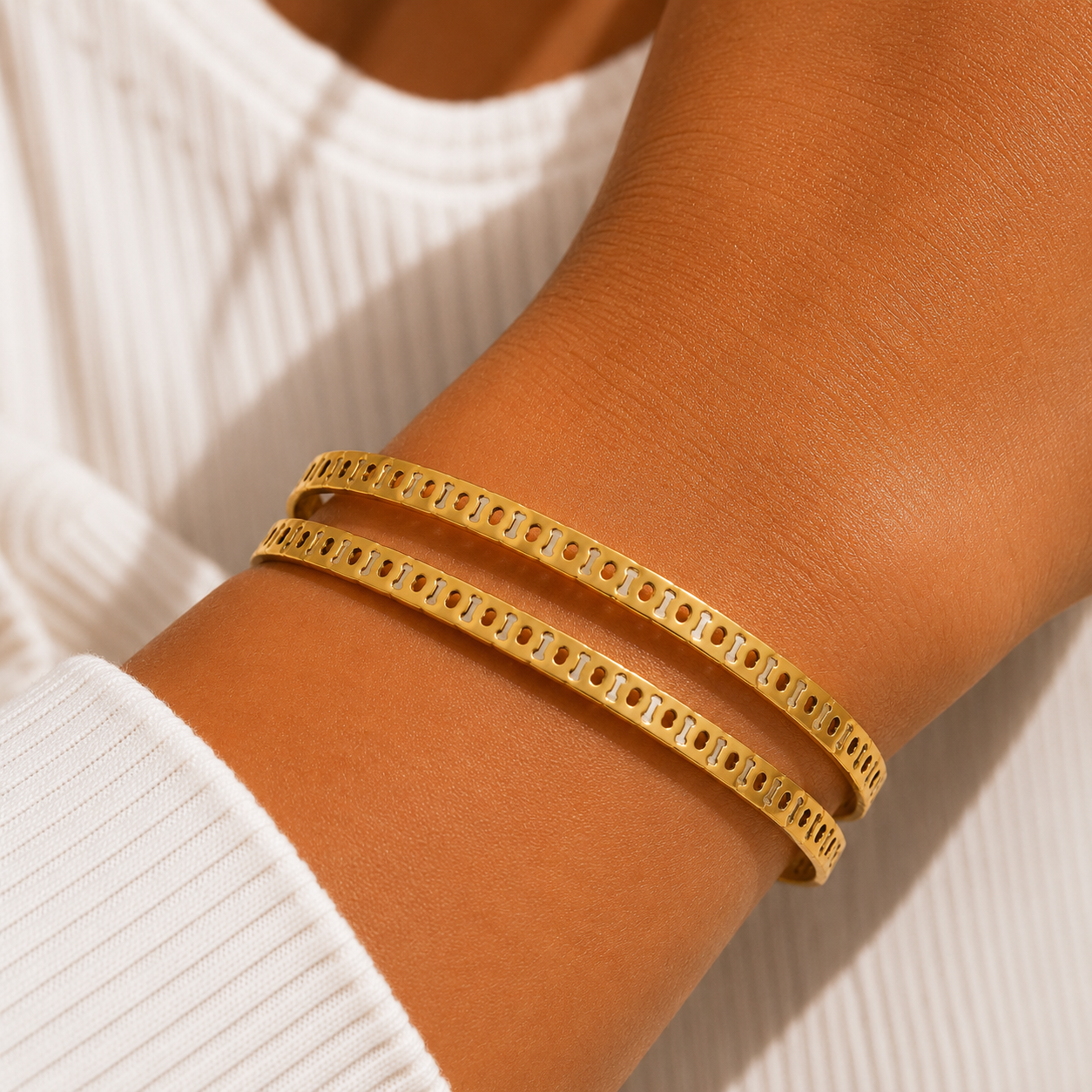 Two gold-plated bangles with a repeating rectangular cutout pattern, worn stacked on a person's wrist over a white ribbed top.