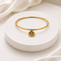 A thin gold bangle bracelet with a small circular pendant featuring a Chinese character for good fortune, displayed on a round white pedestal with soft white fabric in the background.