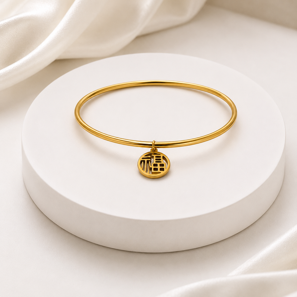 A thin gold bangle bracelet with a small circular pendant featuring a Chinese character for good fortune, displayed on a round white pedestal with soft white fabric in the background.