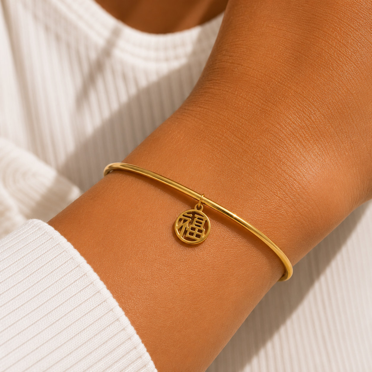 A close-up of a person's wrist wearing a thin gold bangle bracelet with a small circular hanging charm featuring a Chinese character for good fortune, styled with a white ribbed long-sleeve top.