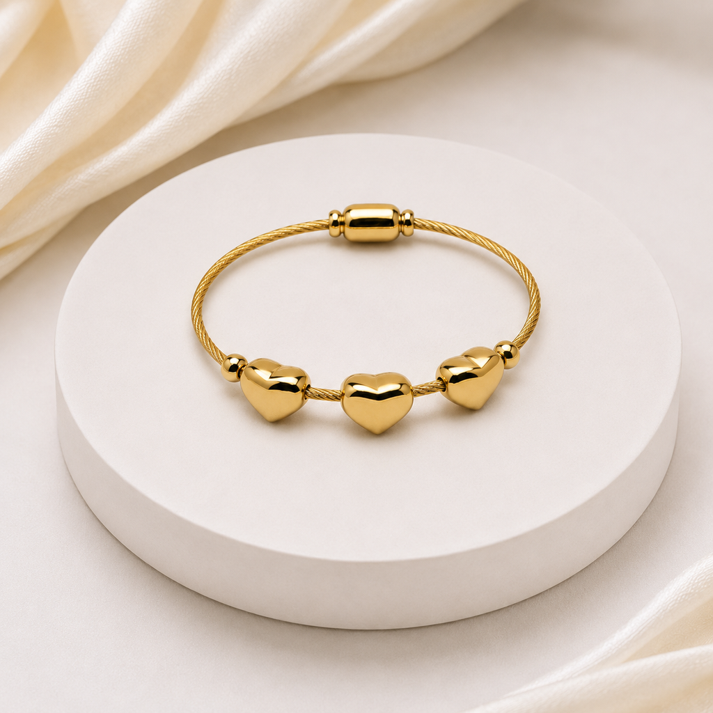 A gold-plated cable bracelet with three polished heart charms, displayed on a round white pedestal with draped cream satin fabric in the background.