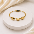 A gold-plated cable bracelet with three polished heart charms, displayed on a round white pedestal with draped cream satin fabric in the background.