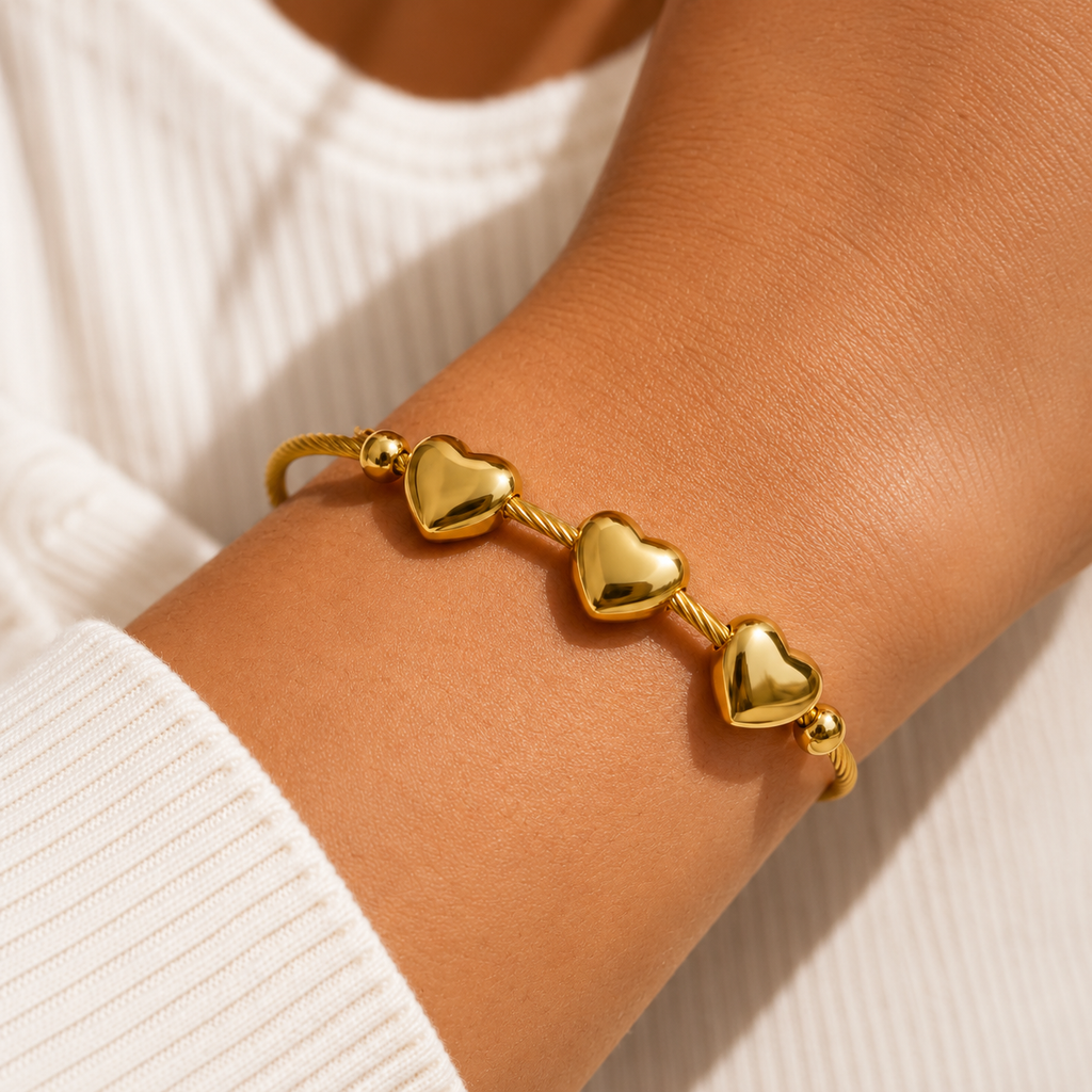 A close-up of a gold heart charm bracelet worn on a person's wrist, paired with a white ribbed knit top.