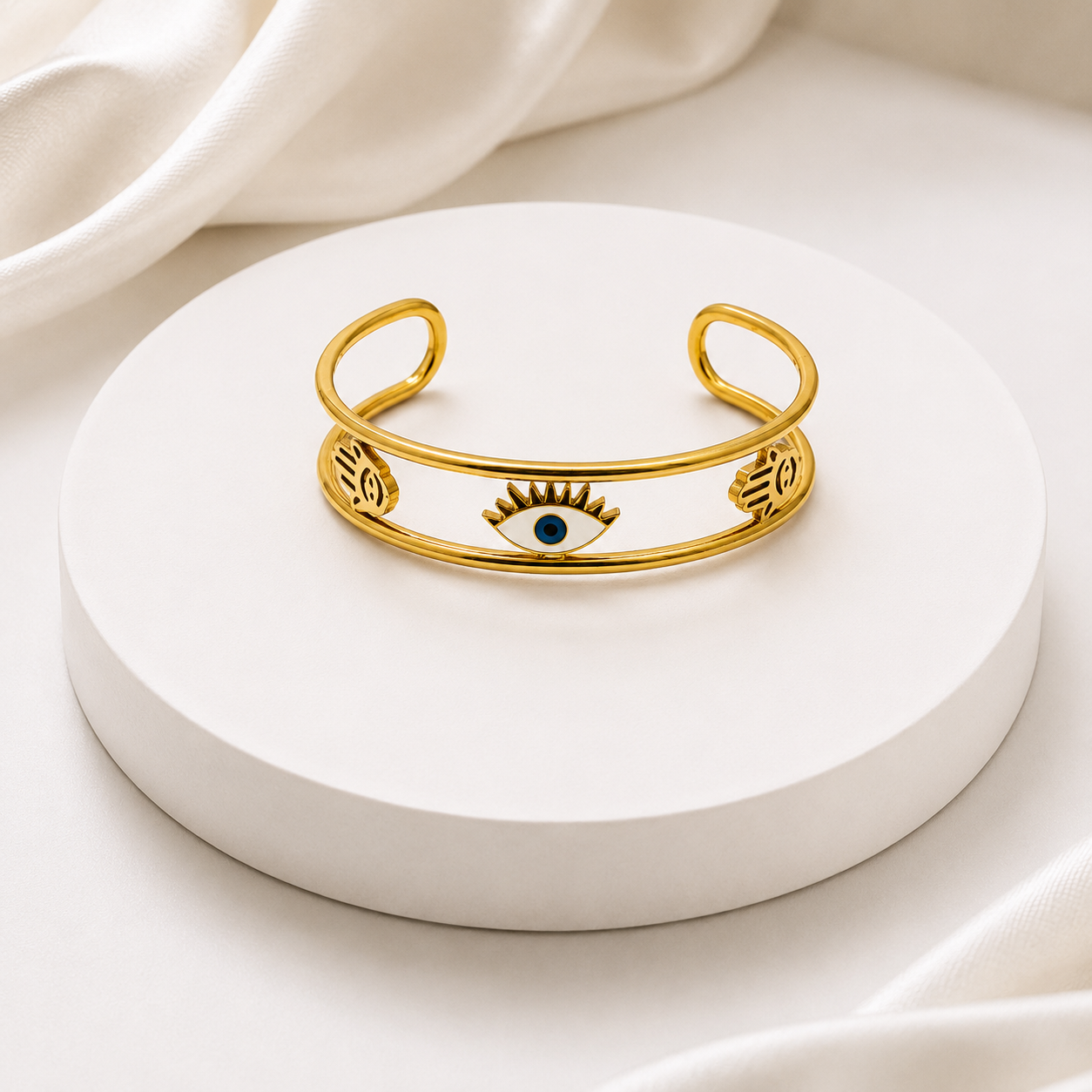 A gold cuff bracelet with an evil eye motif in the center and engraved floral details on the sides, displayed on a round white pedestal.