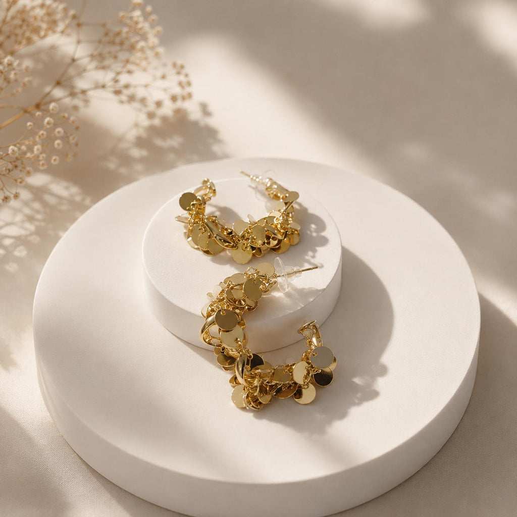 Gold dangling hoop earrings with irregular circular charms, displayed on a round white pedestal with soft shadows and dried floral decor in the background.