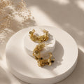 Gold dangling hoop earrings with irregular circular charms, displayed on a round white pedestal with soft shadows and dried floral decor in the background.
