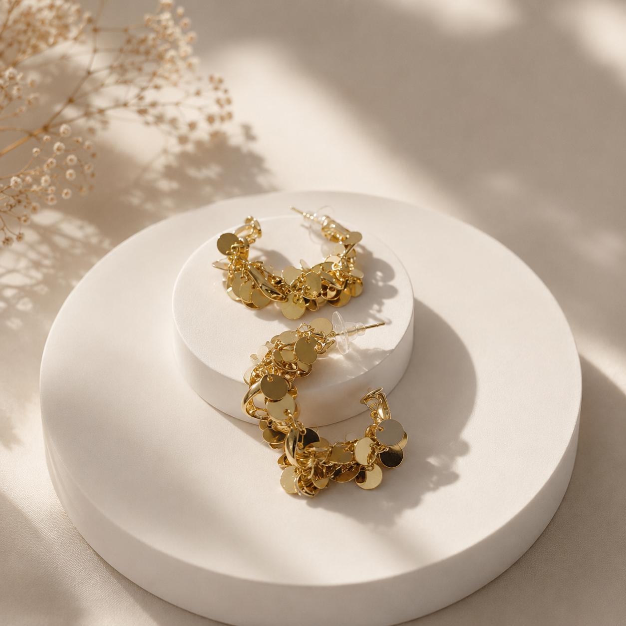 Gold dangling hoop earrings with irregular circular charms, displayed on a round white pedestal with soft shadows and dried floral decor in the background.