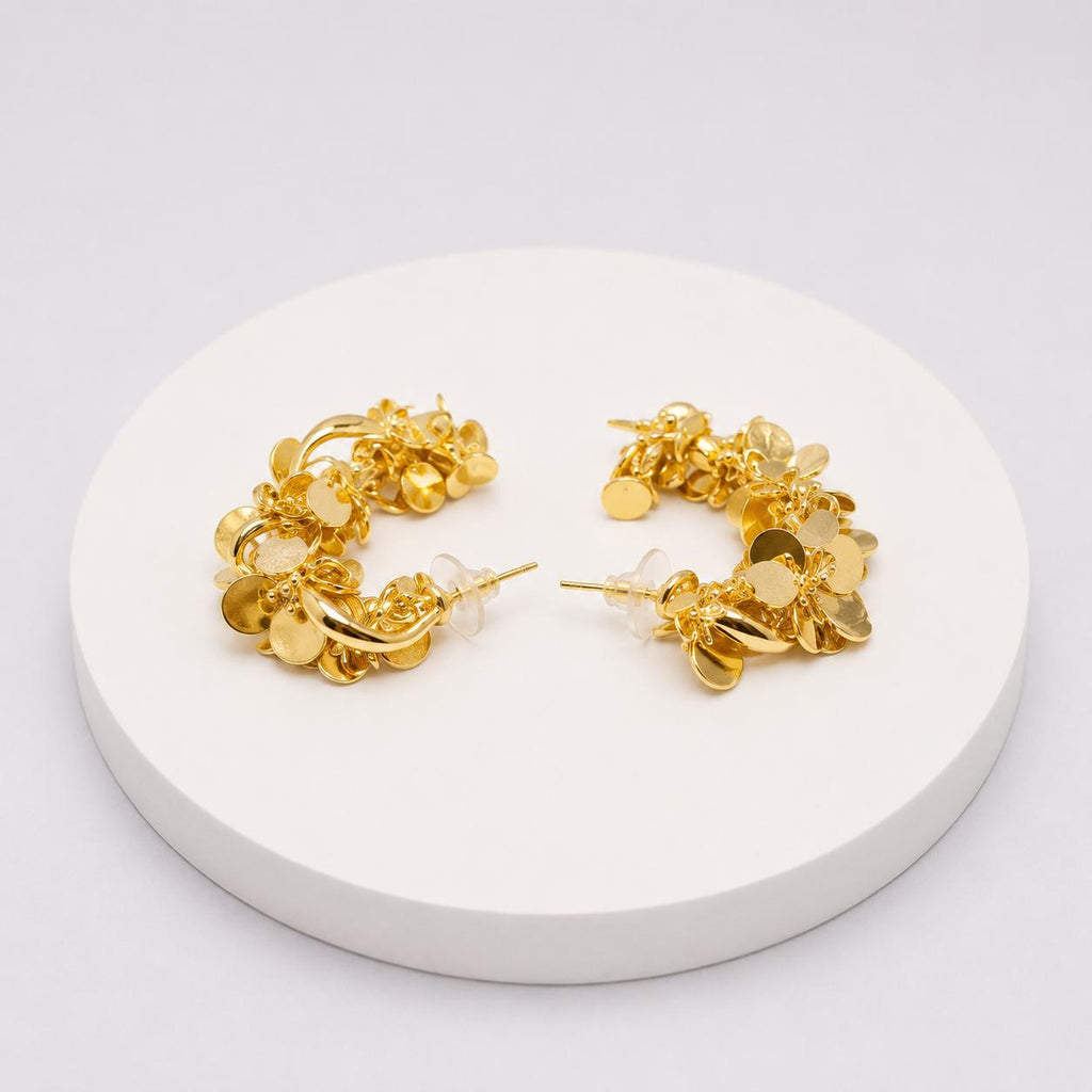 A pair of gold hoop earrings with dangling flower and leaf charms, displayed on a round white pedestal against a light gray background.
