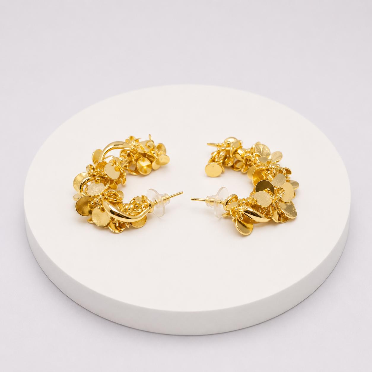 A pair of gold hoop earrings with dangling flower and leaf charms, displayed on a round white pedestal against a light gray background.