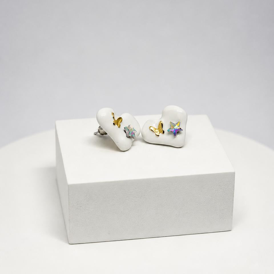 A pair of white heart-shaped stud earrings with small butterfly and flower details, displayed on a white rectangular pedestal against a light gray background.