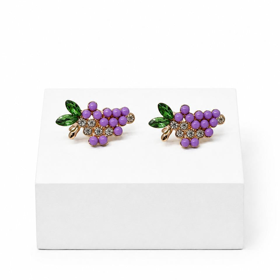 A pair of gold-tone grape cluster stud earrings with purple enamel grapes and green enamel leaves, displayed on a white rectangular block against a white background.