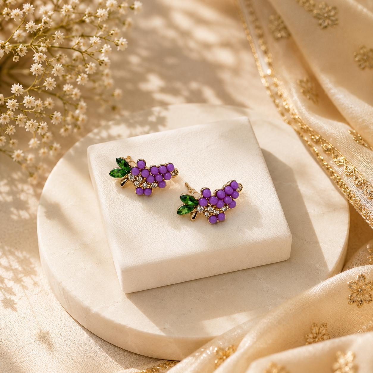 A pair of grape cluster stud earrings with purple enamel grapes, green leaves, and gold-toned settings, displayed on a white jewelry block over a cream satin fabric backdrop.