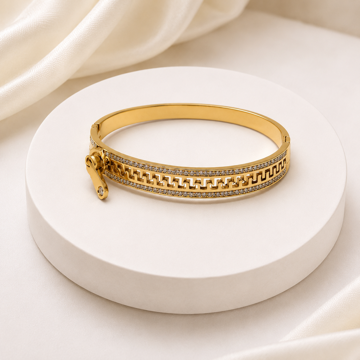 A gold bangle bracelet with a geometric pattern and a row of small clear stones, displayed on a round white pedestal with draped white fabric in the background.